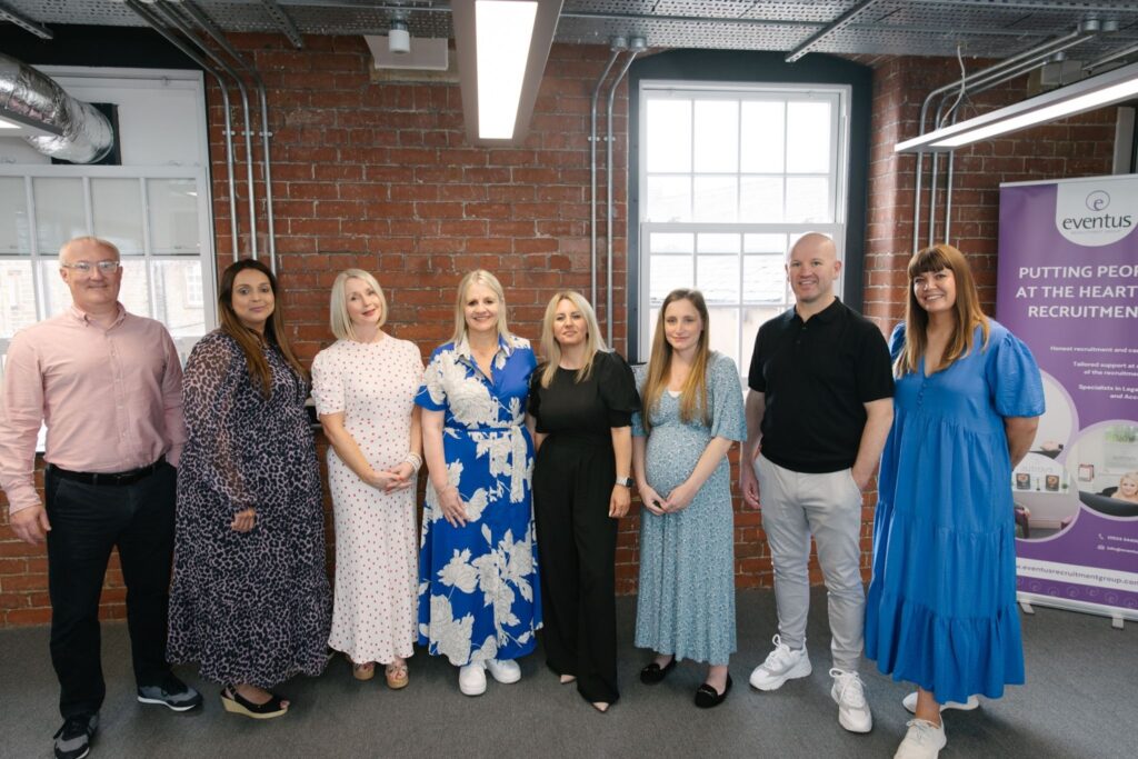 Image shows the full team of Recruitment Consultants at Eventus Recruitment Group in the office, standing by a red brick wall with a banner in the background.